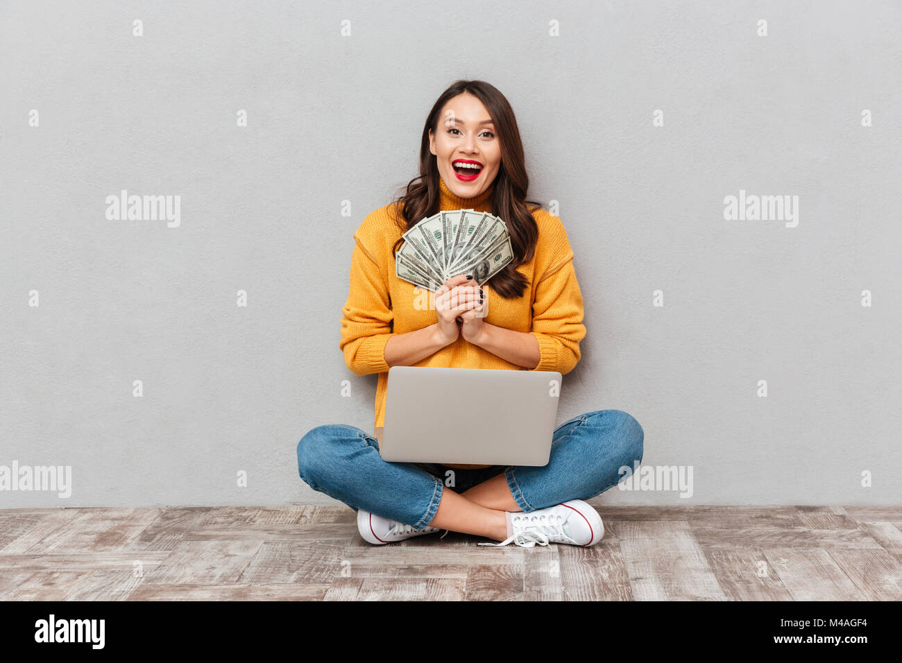 Cheerful brunette woman in sweater assis sur le plancher avec un ordinateur portable et la tenue de l'argent tout en se réjouit et regardant la caméra sur le dos gris Banque D'Images