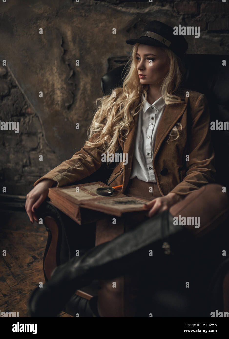 Jeune femme pensive à l'image de Sherlock Holmes est assis dans un fauteuil et est titulaire d'ancien photoalbum dans ses mains avec loupe sur fond de murs en briques anciennes Banque D'Images