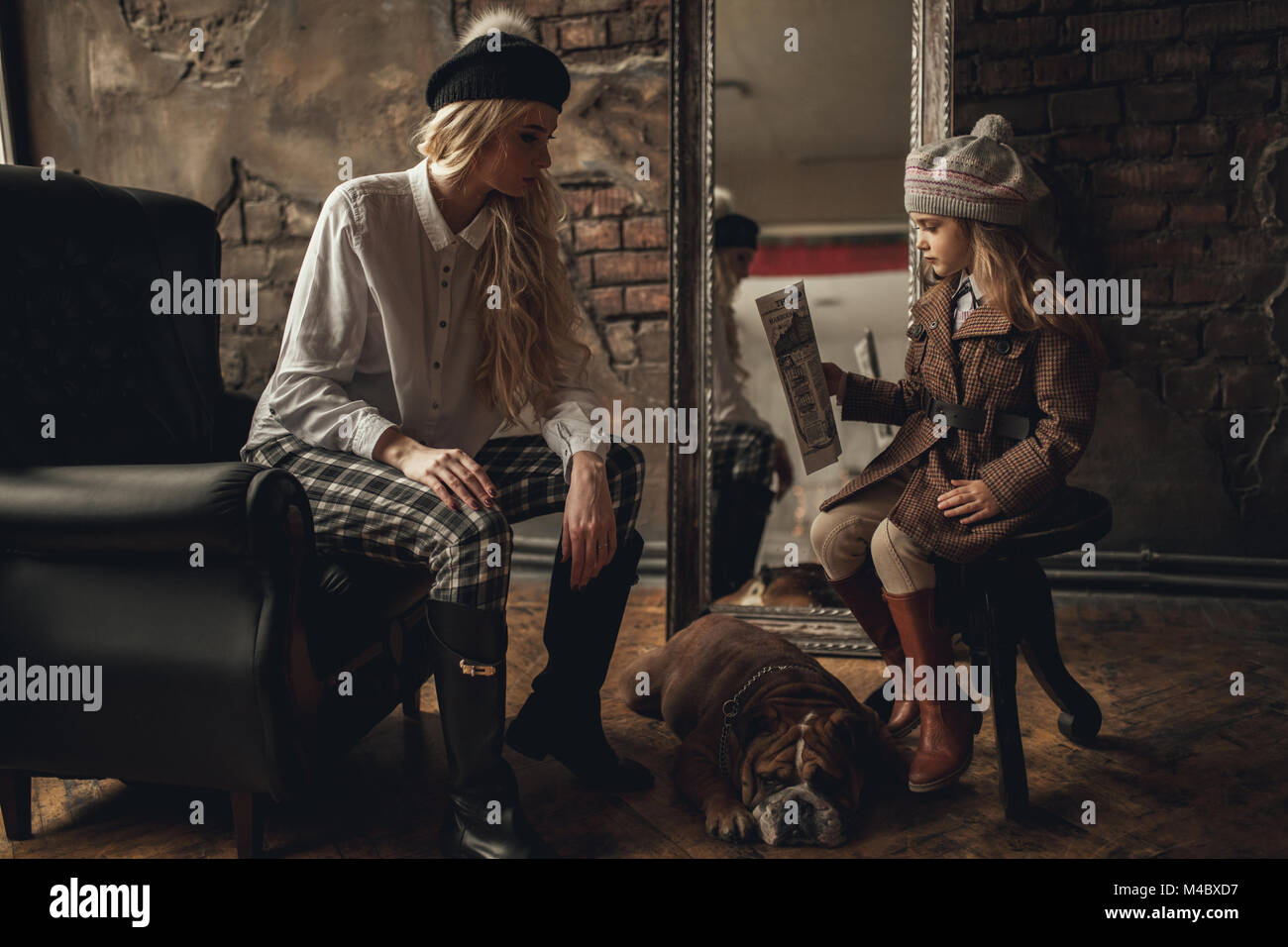 Enfant fille avec femme dans l'image de Sherlock Holmes s'asseoir dans un fauteuil et lire à côté de journal bulldog Anglais sur fond de murs en briques anciennes, miroir Banque D'Images