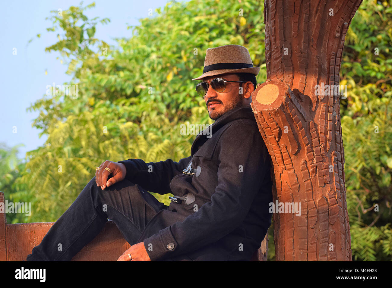 Homme dans un chapeau et blouson noir assis contre un tronc d'arbre, Pune, Maharashtra. Banque D'Images