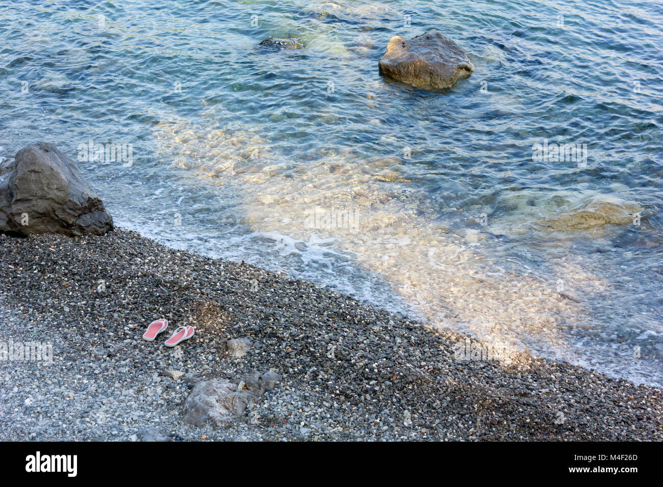 Bord de l'eau d'une plage rocheuse avec une paire de tongs roses sur les galets. Banque D'Images