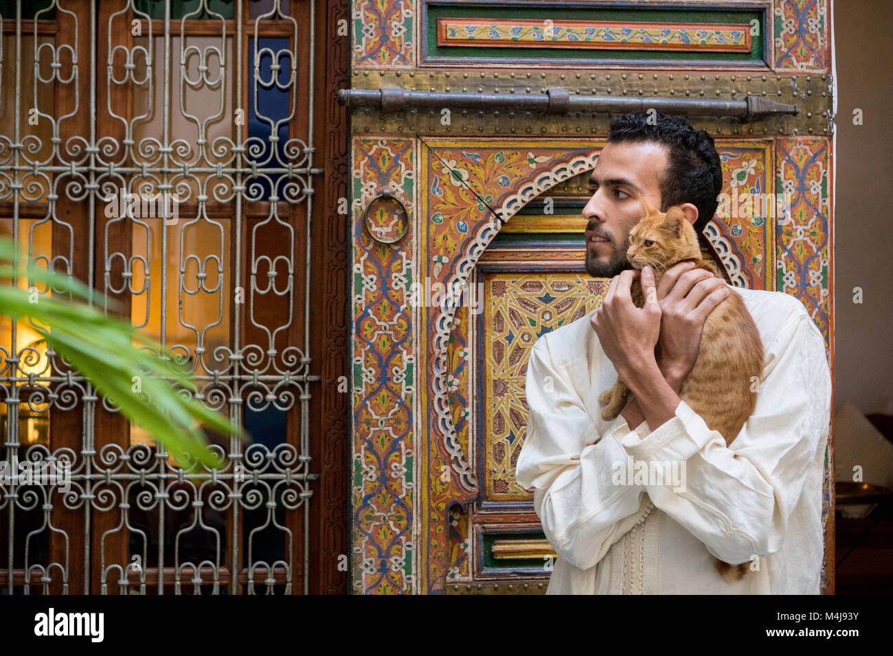Jeune homme en costume traditionnel musulman tenant un chat jaune dans le style marocain de la température ambiante en face d'un mur décoré Banque D'Images
