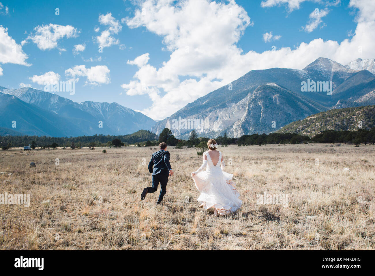 Mariée et le marié s'enfuyant avec dos à l'appareil photo dans un champ avec un décor de montagnes sur un jour d'été parfait avec ciel bleu et nuages blancs grand Banque D'Images