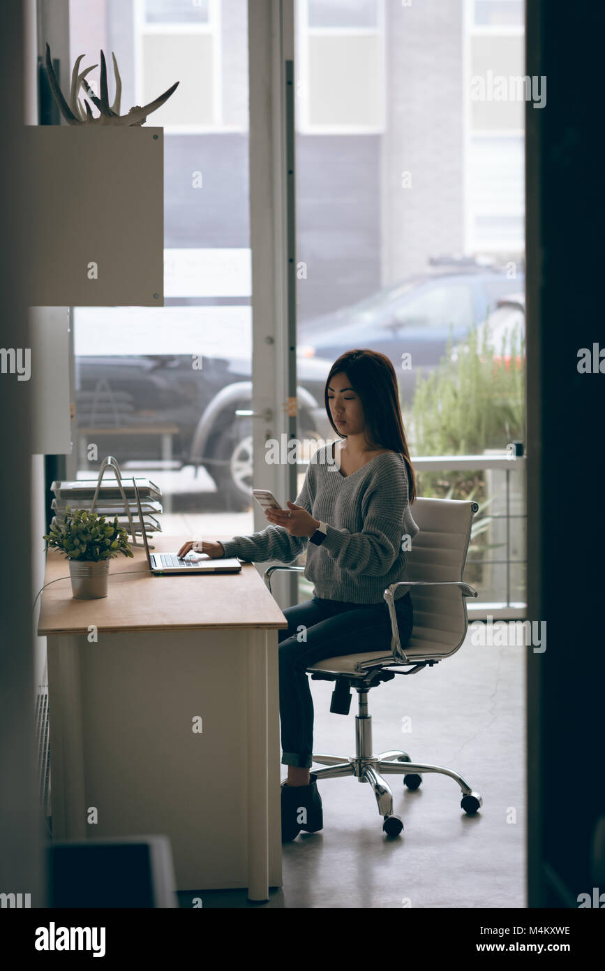 Female executive using mobile phone while working on laptop at desk Banque D'Images