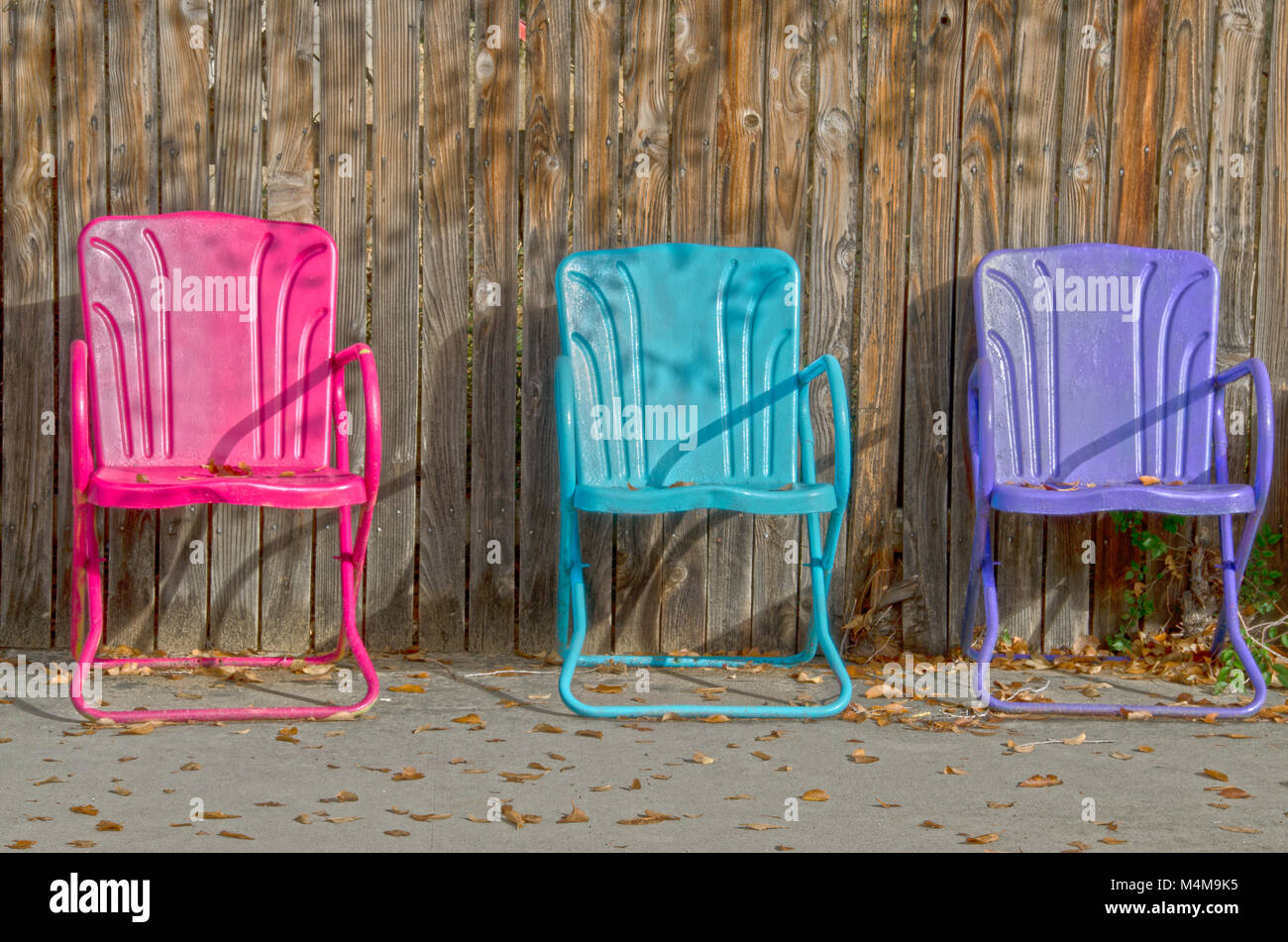 Trois chaises de jardin coloré vide s'asseoir contre une clôture en bois, avec les feuilles d'automne dispersés parmi eux. Banque D'Images