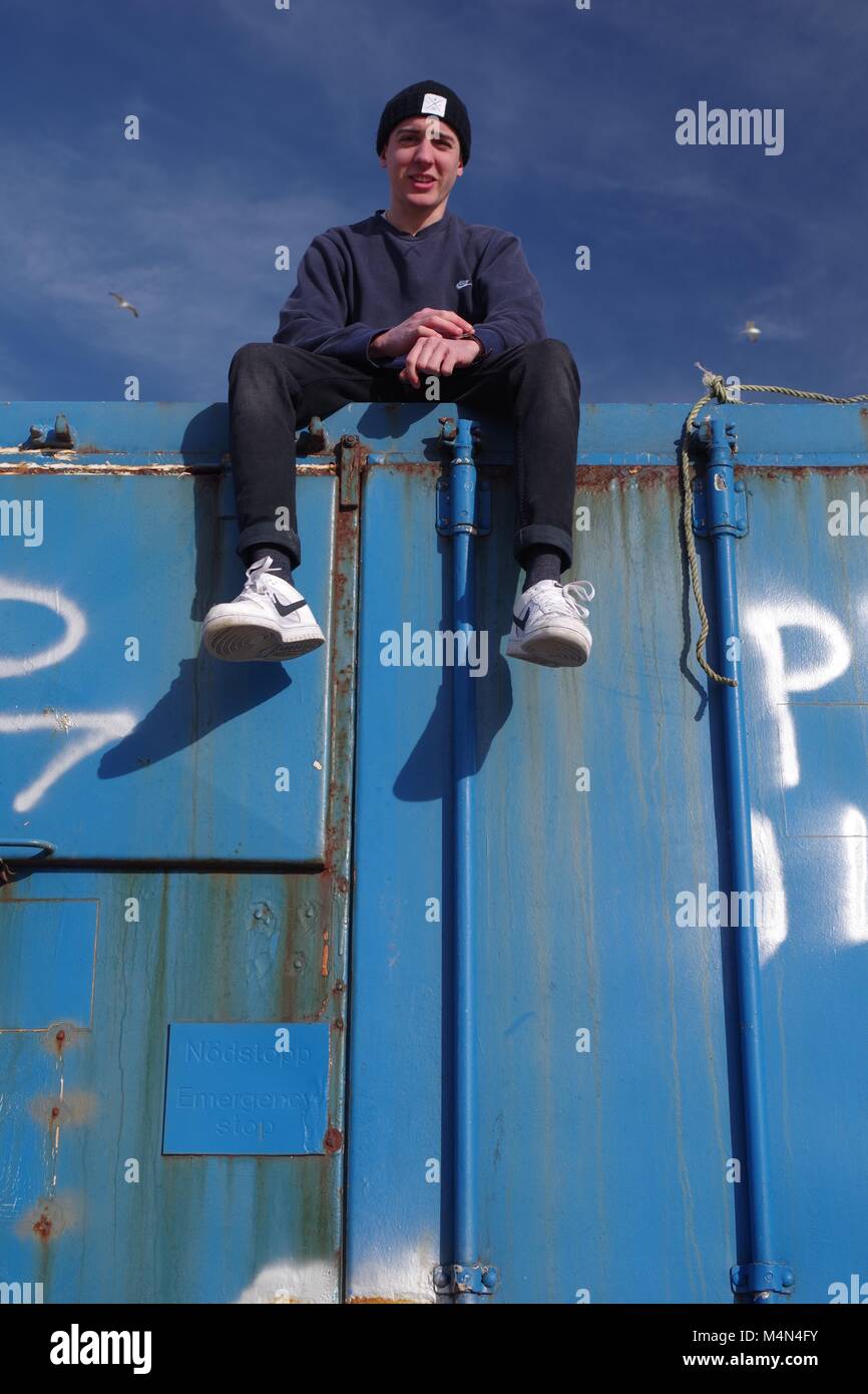 Jeune homme assis au sommet d'un ancien bleu contenant d'expédition au port Bay Cruden, Aberdeenshire, Scotland, UK. L'hiver, en 2018. Banque D'Images