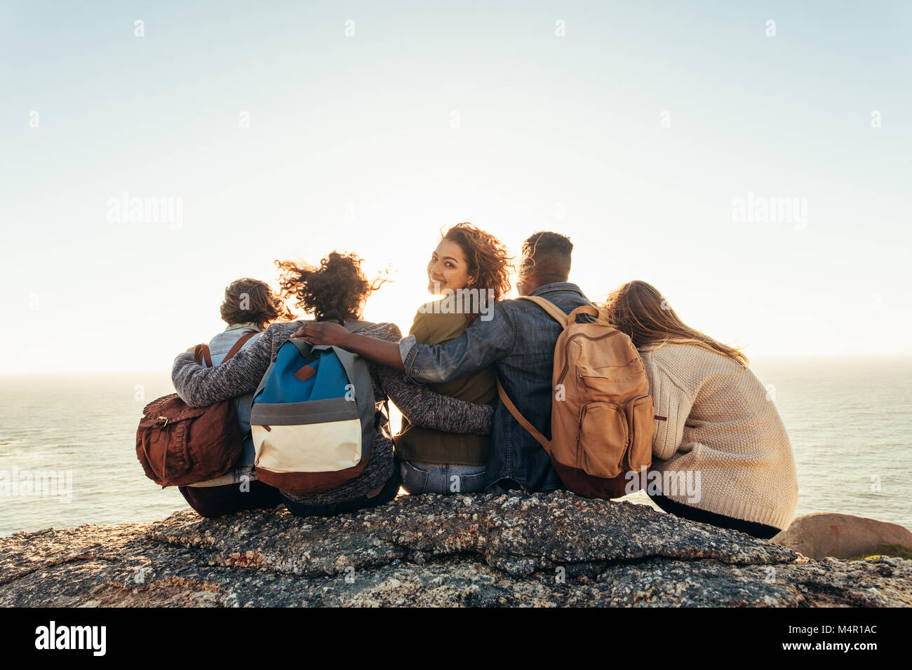 Groupe de jeunes hommes et femmes assis en montagne et admirer le coucher du soleil. Jeune femme avec des amis passer une journée à l'extérieur. Banque D'Images