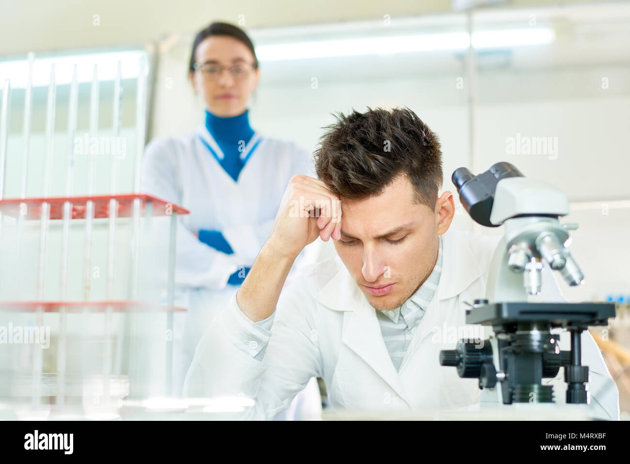 Jeune chercheur concentré wearing white coat penche la tête sur la main tout en siégeant au bureau, sa jolie collègue derrière lui, de l'intérieur Banque D'Images