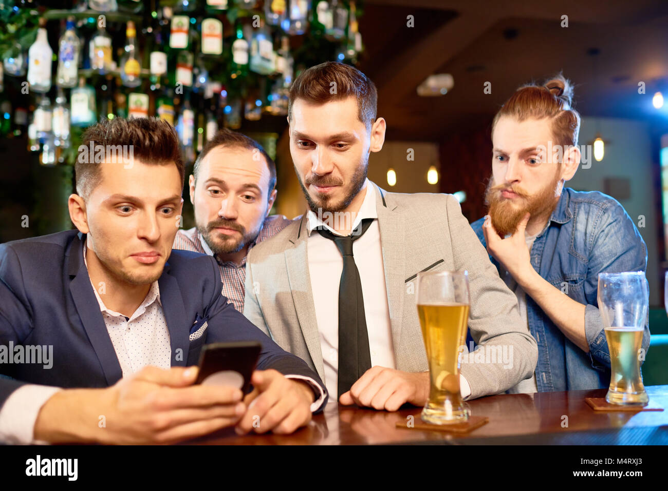 Groupe d'hommes barbus surpris rassemblés en bar, boire de la bière et la lecture des nouvelles avec l'aide du smartphone Blackberry Banque D'Images