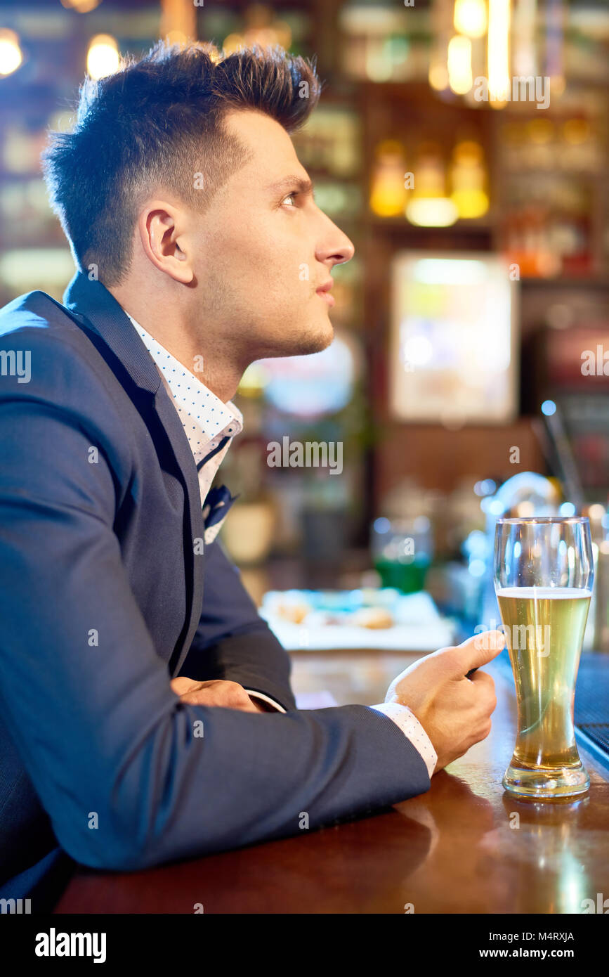 Voir le profil de beau jeune homme portant un costume élégant assis au  comptoir du bar après une dure journée de travail, de boire une bière et  regarder des match de rugby