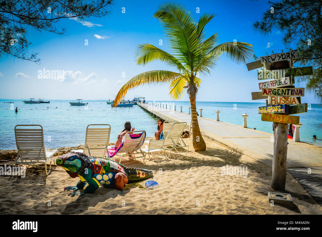 Destinations internationales signpost au quai à côté de chaises longues et de palmiers sur la plage avec l'iguane bleu statue au Club Rum Point, Grand Cayman. Banque D'Images