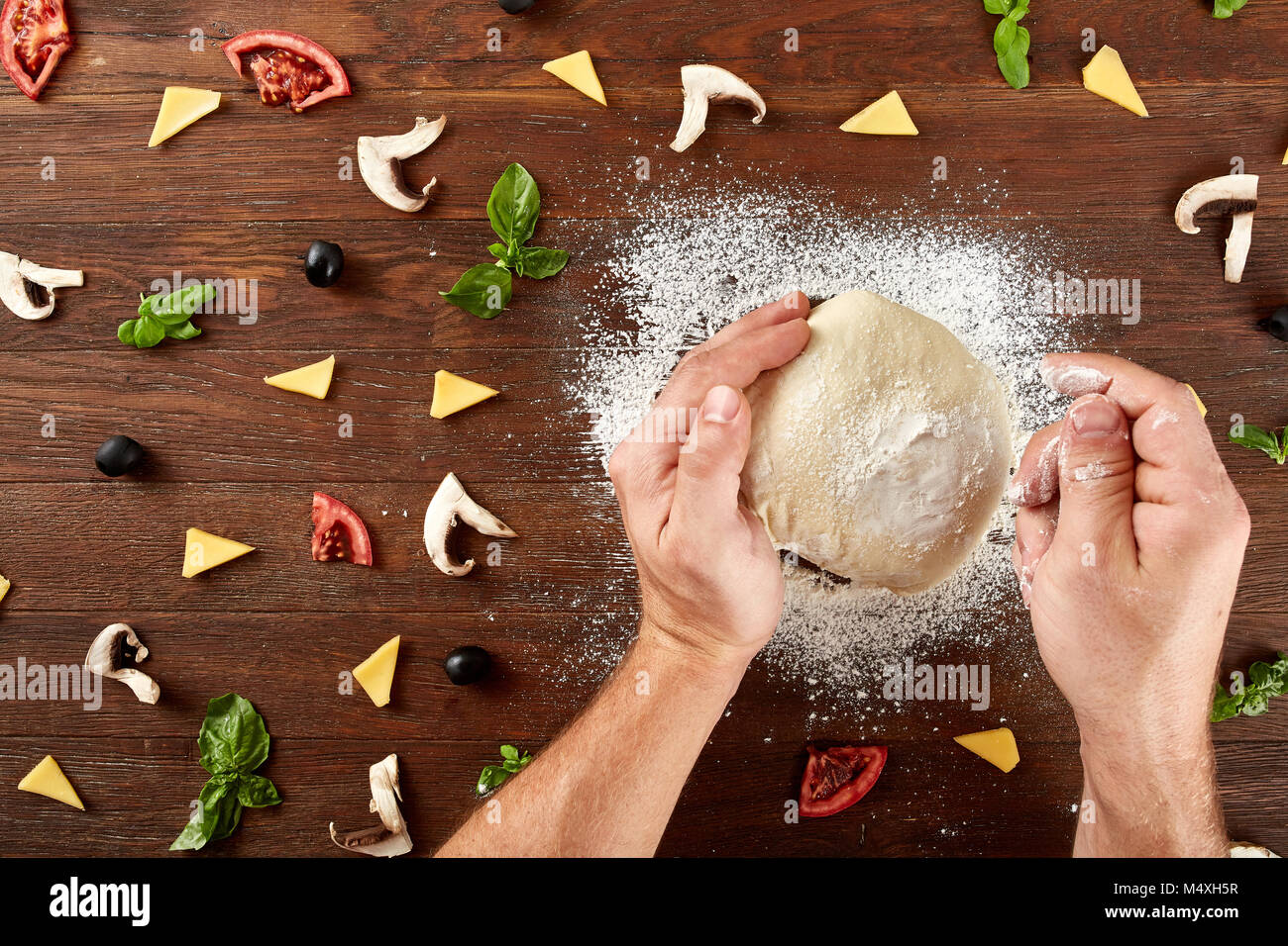 Close-up la main d'un pétrir et façonner la pâte Baker pour la pizza sur une table en bois brun foncé, encadré par quelques morceaux de tomates, champignons, fromage, Banque D'Images