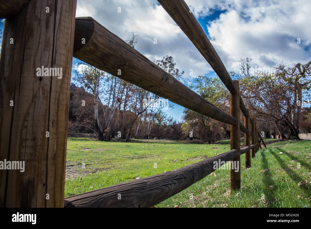 Clôture en bois rustique coulée le long de l'ombre après vert champ prairie sous ciel nuageux. Banque D'Images