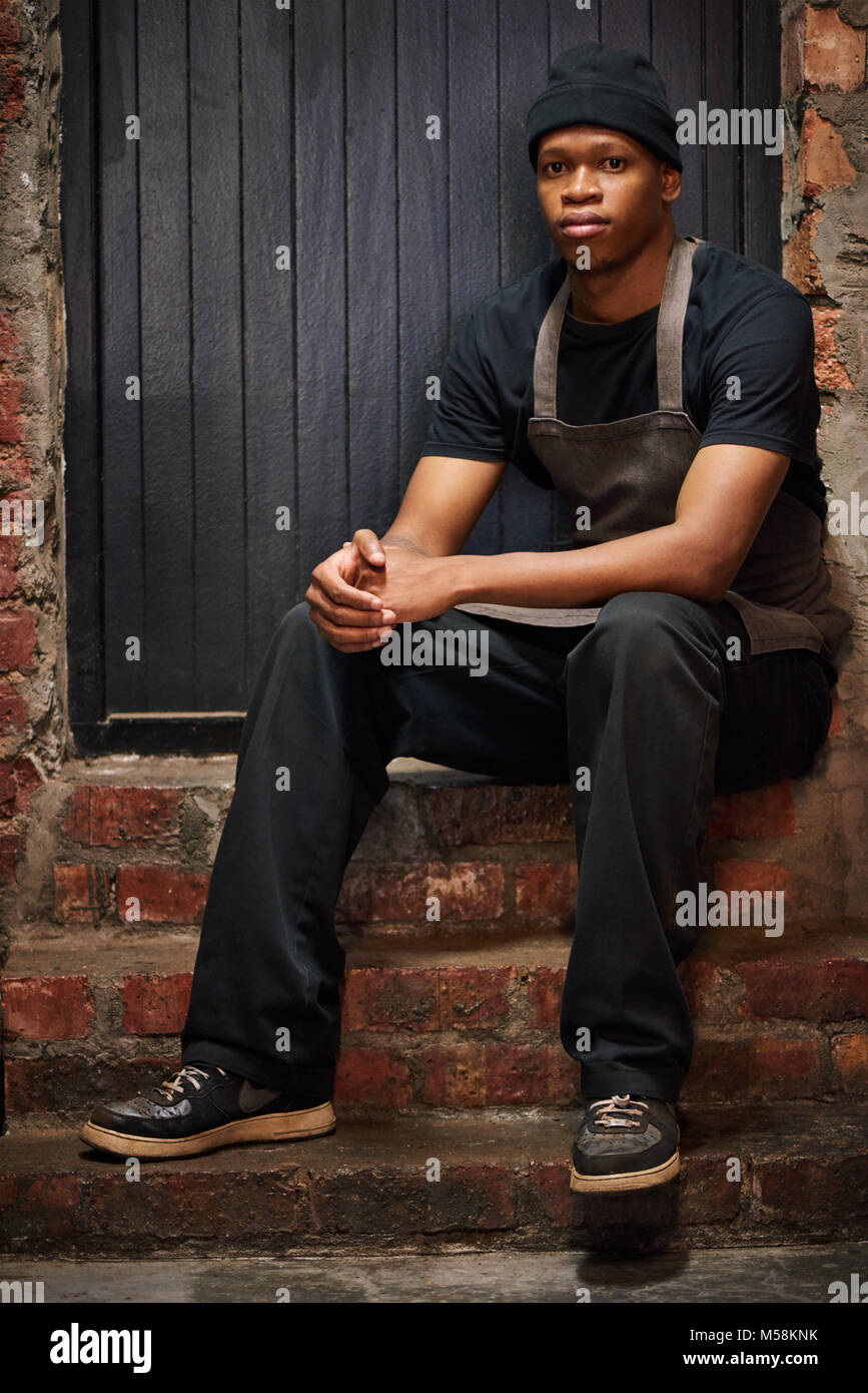 Portrait de barista africain portant un tablier marron clair et les vêtements noirs ainsi qu'un bonnet noir, avec un mur de briques et de faire face à l'ajout de certaines mesures con Banque D'Images