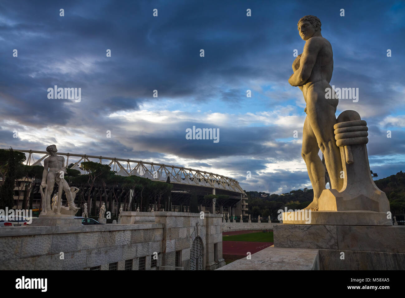 Des statues représentant des athlètes au Stadio dei Marmi à Rome. Dans l'arrière vous pouvez voir le stade olympique. La scène est éclairée par la lumière et le coucher du soleil spectaculaire Banque D'Images