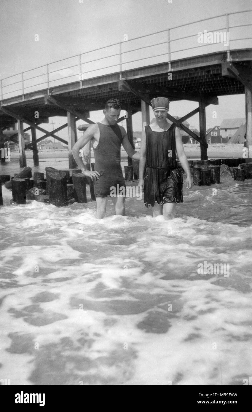 Un jeune couple se trouve dans le surf sur la promenade tout en nageant dans l'océan Atlantique, ca. 1920. Banque D'Images
