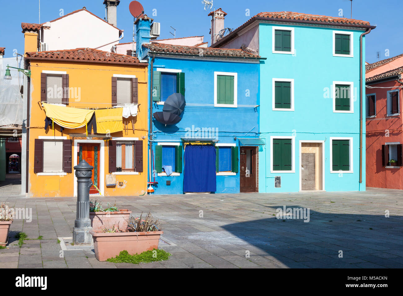 L'île de Burano, Venise, Vénétie, Italie. Maisons colorées de bleu et jaune autour d'une petite place avec une fontaine d'eau . Le lavage et de parasols sur la pendaison Banque D'Images