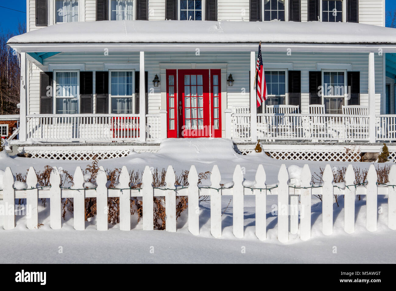 New Englander traditionnelle maison avec porte avant rouge, drapeau américain, clôture et clôture autour de porche en hiver avec de la neige à Franconia, NH, USA. Banque D'Images