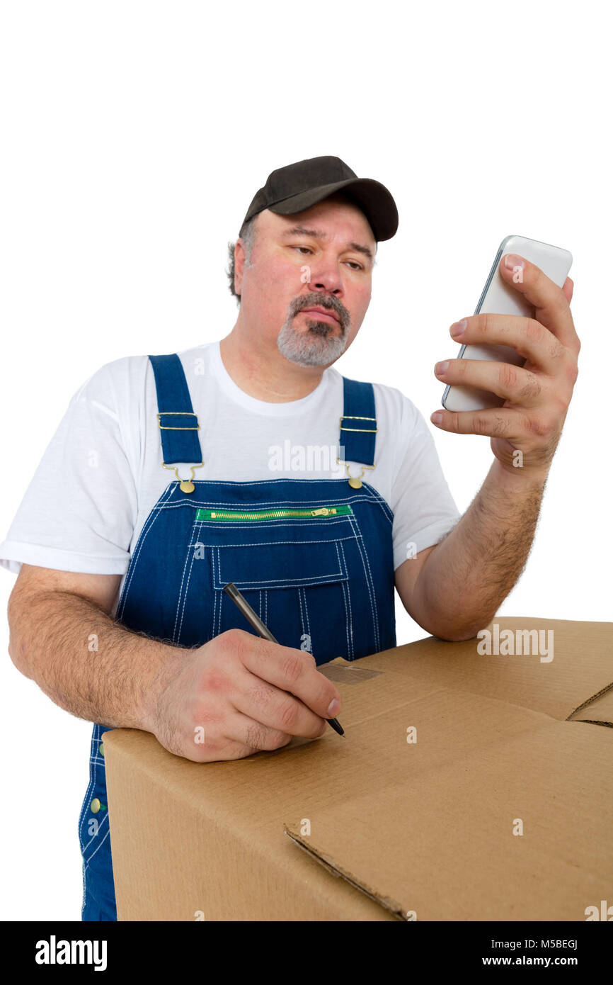 Travailleur en bleu de travail lecture d'un message texte sur son smartphone qu'il note les instructions pour une grande boîte en carton qu'il s'appuie sur isolated on white Banque D'Images