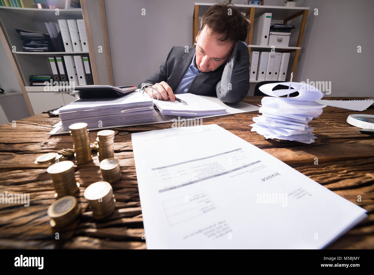 Stressed Businessman working in office avec facture et des pièces d'or empilés sur un bureau en bois Banque D'Images