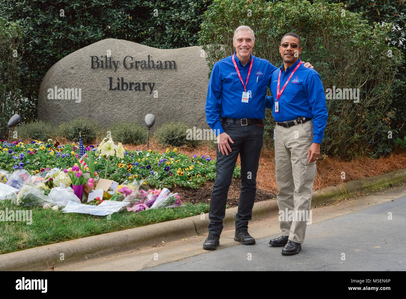 Charlotte, NC, USA. 22 Février, 2018. Billy Graham Evangelistic Association (BGEA) les employés se réunissent à l'avant portes de la Billy Graham Library pour aider bienfaiteurs apportant fleurs et cadeaux en l'honneur du pasteur qui est mort 21 février à l'âge de 99 ans. L'aumônier BGA, Terry Sartain, et BGEA Manager de l'intervention d'urgence et de la logistique, Kevin Williams, accueillir les visiteurs aux portes et de rappeler aux spectateurs que "Jésus vous aime." Bien qu'attristé par le révérend Graham's passant, ce mémorial est également considérée comme une semaine de célébration de la vie de Graham Rev.. Credit : Château Light Images/Alamy Live News. Banque D'Images