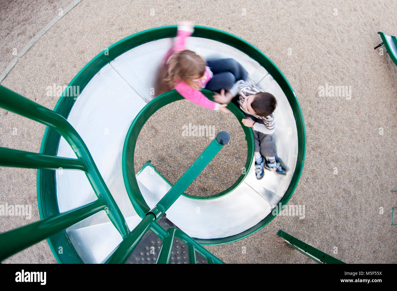 En regardant un jeune garçon et fille glissant sur une spirale metal diapositive d'une aire de jeux, flou de mouvement, perspective fisheye. Banque D'Images