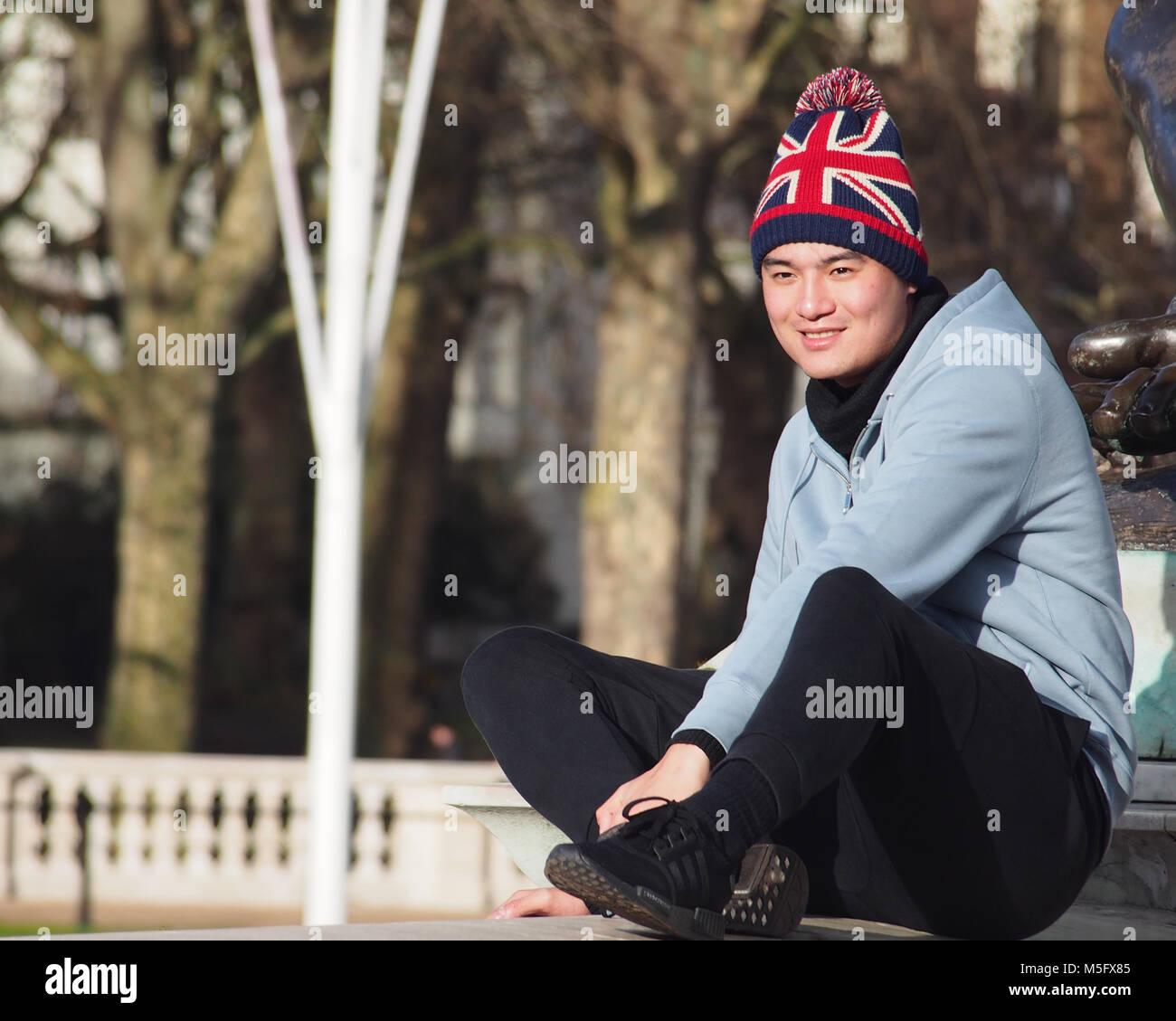 Jeune homme, touristiques, assis jambes croisées sur un mur dans l'avant-cour du palais de Buckingham portant un drapeau de l'Union bobble hat dans winter sunshine Banque D'Images