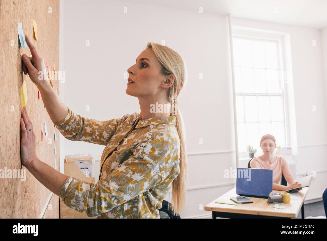 Travailleur féminin pointe vers une note sur un panneau de liège et est d'avoir une discussion avec un de ses collègues, qui est assis à un bureau à l'arrière-plan. Banque D'Images