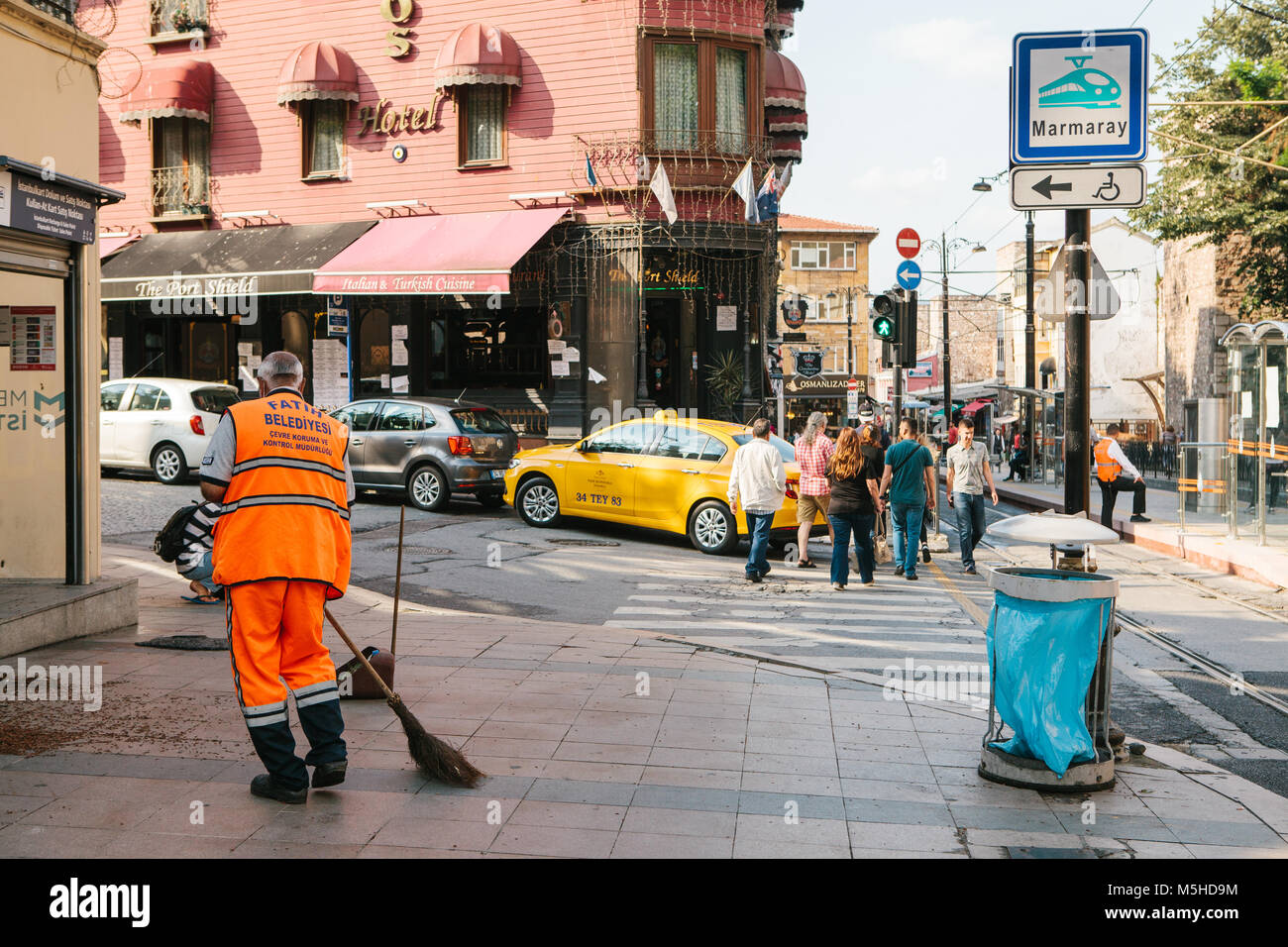 Concierge dans uniforme orange lumineux balayant la tuile sur la rue dans le quartier de Sultanahmet à Istanbul, Turquie. Les piétons qui traversent la route dans le bac Banque D'Images