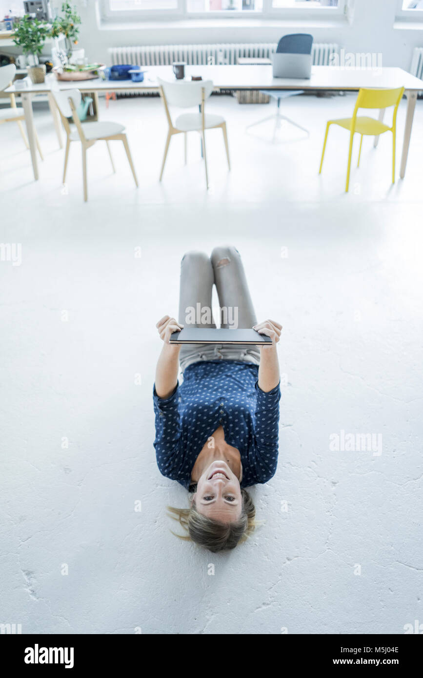 Portrait of laughing young woman with tablet gisant sur le sol dans un loft Banque D'Images