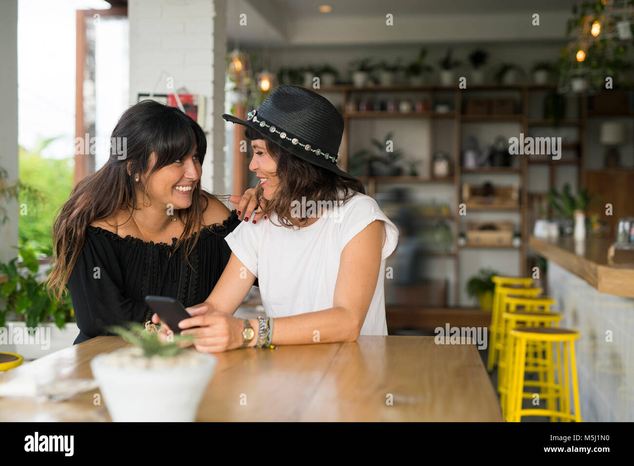 Deux femmes friends smiling at each other in cafe with smartphone Banque D'Images