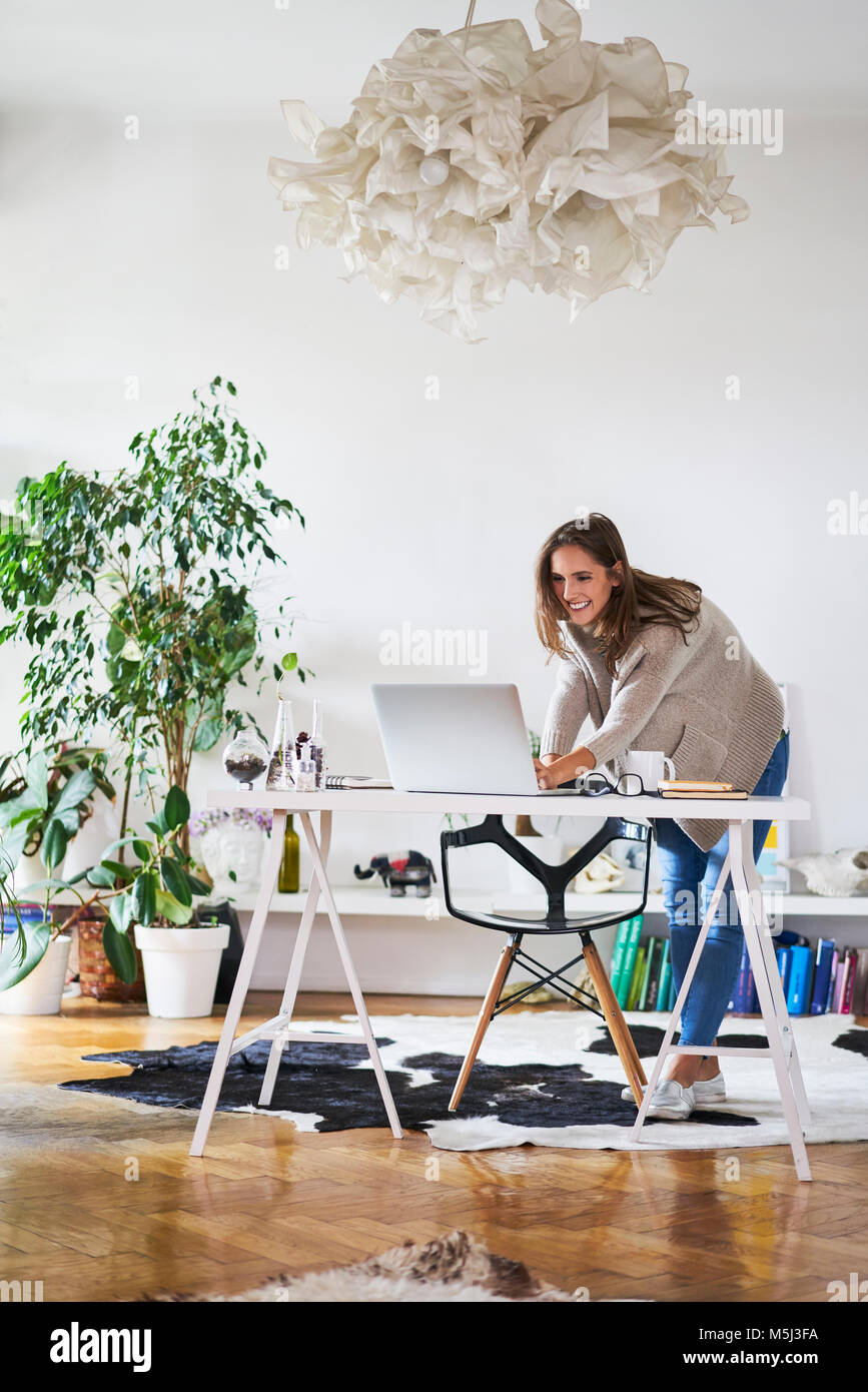Smiling young woman at home using laptop on desk Banque D'Images