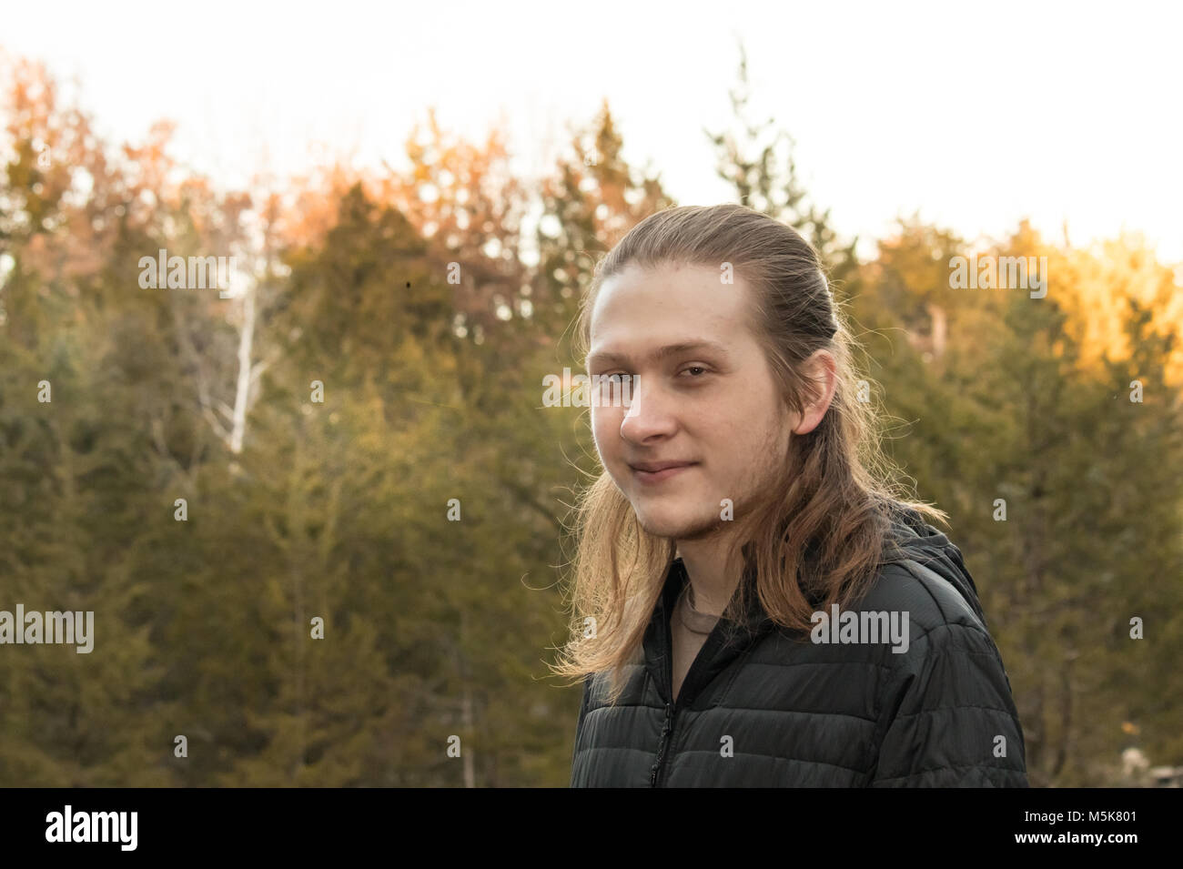 Portrait d'un jeune homme aux cheveux longs avec des arbres en arrière-plan ; la fin de l'après-midi, coucher du soleil Banque D'Images
