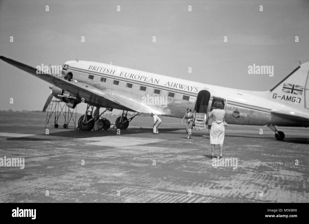 Un Douglas DC-3 Dakota, ou C-47, G-AMGD, appartenant à BEA, British European Airways, à un aéroport inconnu en 1960. Une hôtesse de l'air peut être vu en train de renoncer à l'avion, qui a sa porte arrière ouverte, tandis qu'un ingénieur est sous l'aile, et une femme prend une photo de l'avion. Banque D'Images