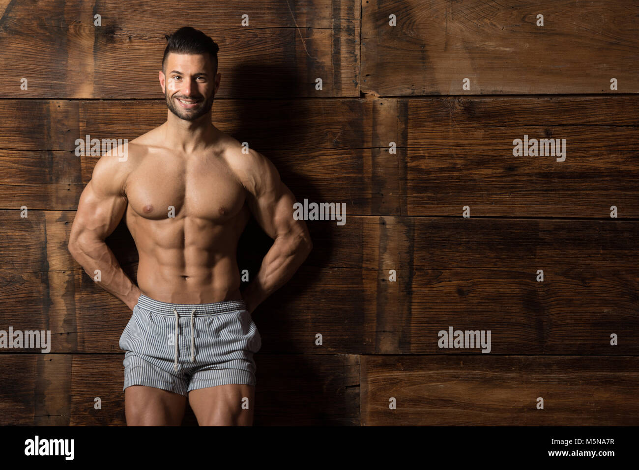 Jeune homme sain Soyons forts contre un mur en bois et Flexing muscles tout en portant des shorts Sports - Athlétisme remise en forme musculaire Bodybuilder Modèle Banque D'Images