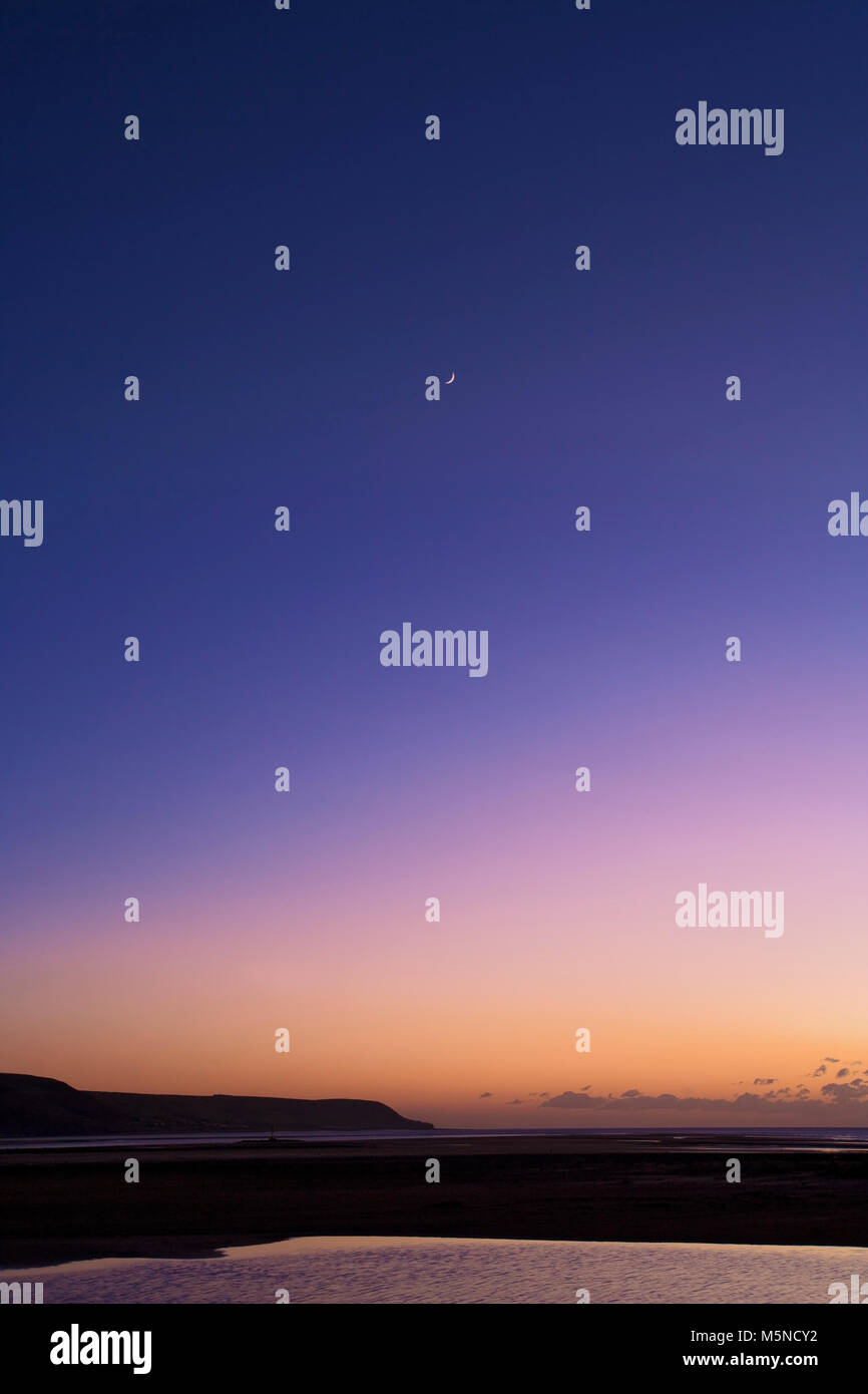 Une nouvelle lune ajoute un point de convergence pour un coucher de soleil sur Barmouth Bay, Pays de Galles. Banque D'Images