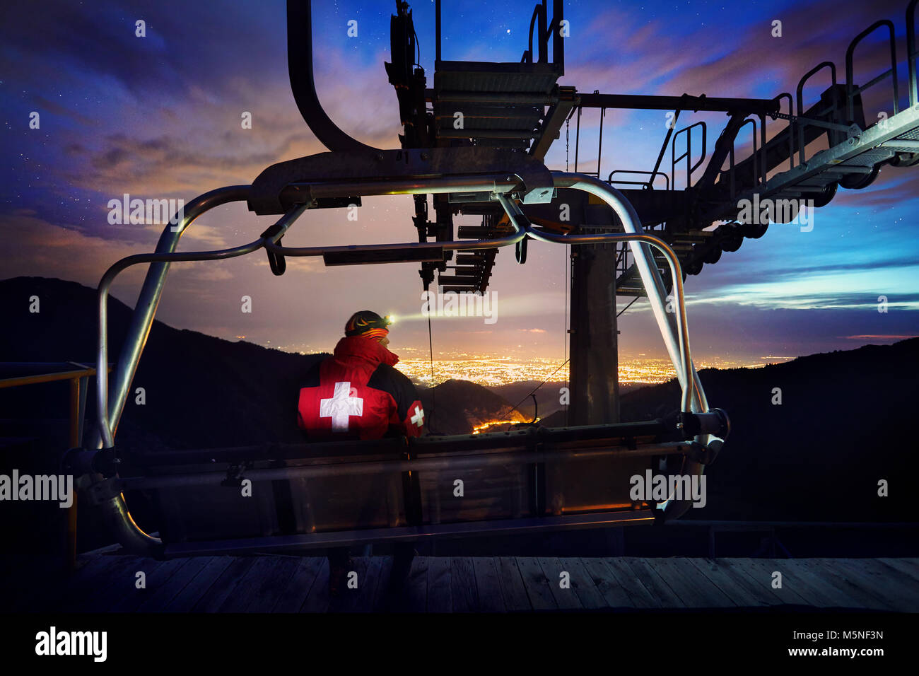 Lifeguard en veste rouge avec croix médicale de l'emplacement sur la chaise en gare du téléphérique avec vue de nuit ville lumière dans le Mountain Ski Resort Banque D'Images