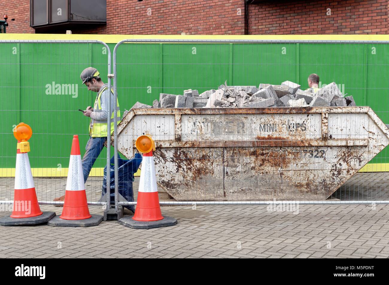 Builder dans un casque et veste haute à savoir l'utilisation d'un téléphone mobile s'appuyant sur un saut rempli de décombres England UK Banque D'Images