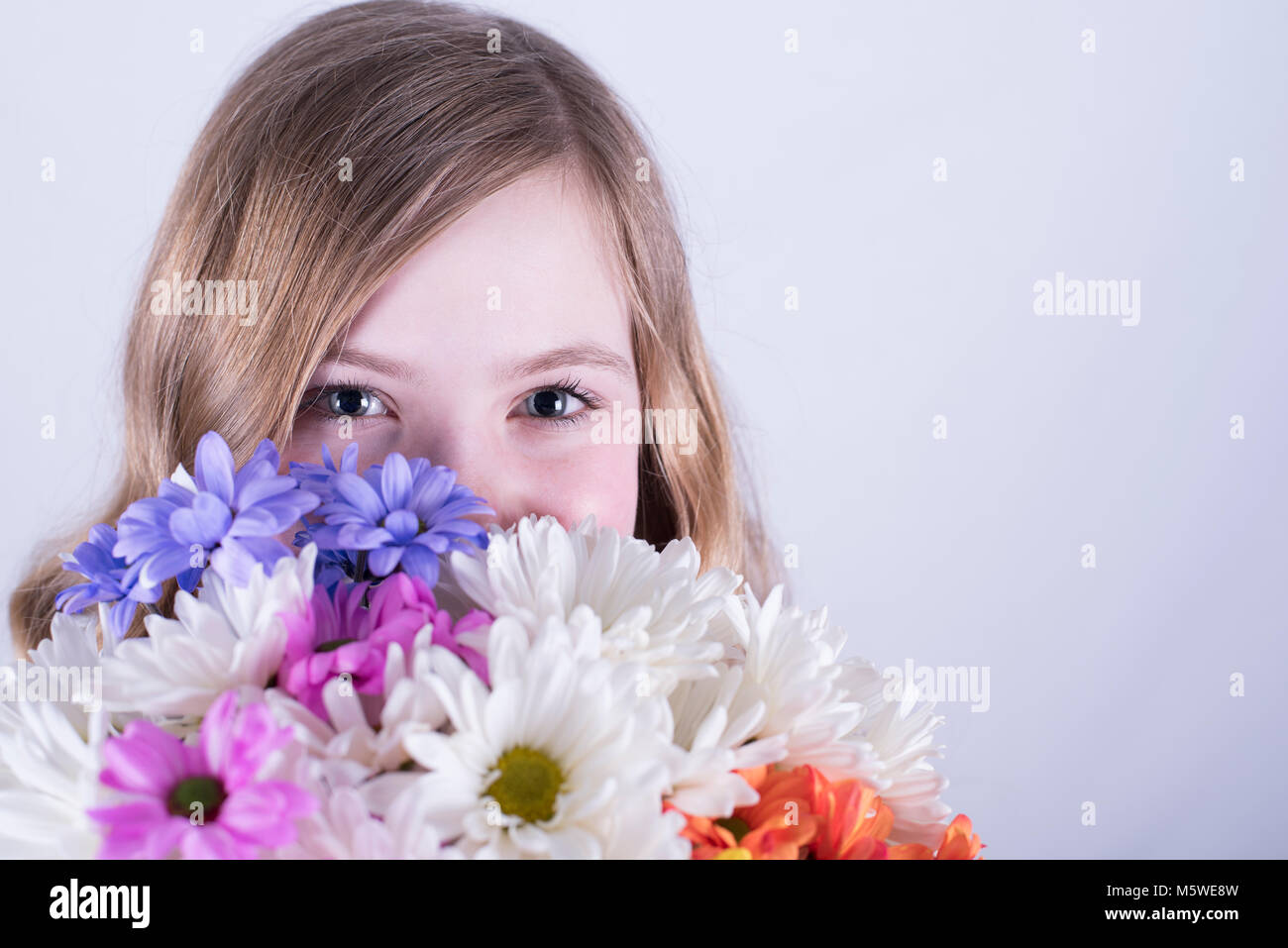 Head shot of 12-year-old fille avec de longs cheveux blond sale, holding bouquet de marguerites colorées sur la face inférieure, la bouche couverte, fond blanc Banque D'Images
