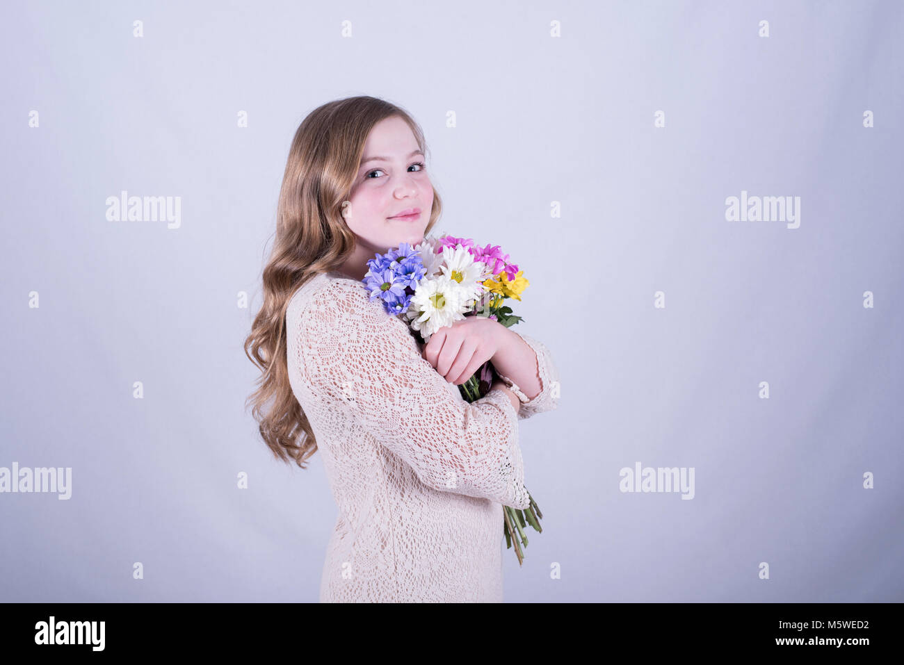 Smiling 12-year-old fille avec de longs cheveux blonds, sales et incliné d'un bouquet de marguerites colorées serrant contre fond blanc Banque D'Images