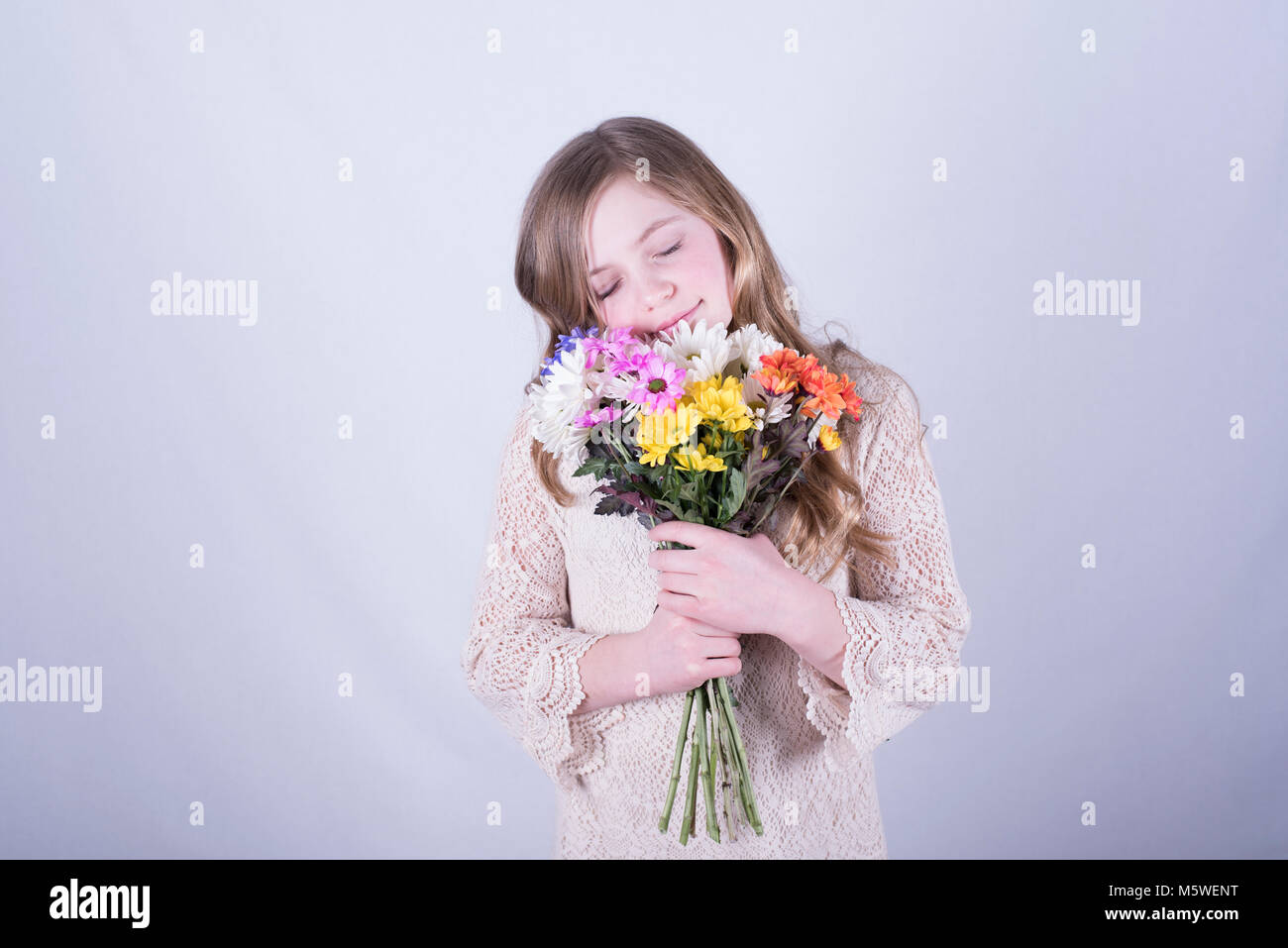 Smiling 12-year-old fille avec de longs cheveux blond sale, serrant le bouquet de marguerites colorées avec les yeux fermés contre fond blanc Banque D'Images