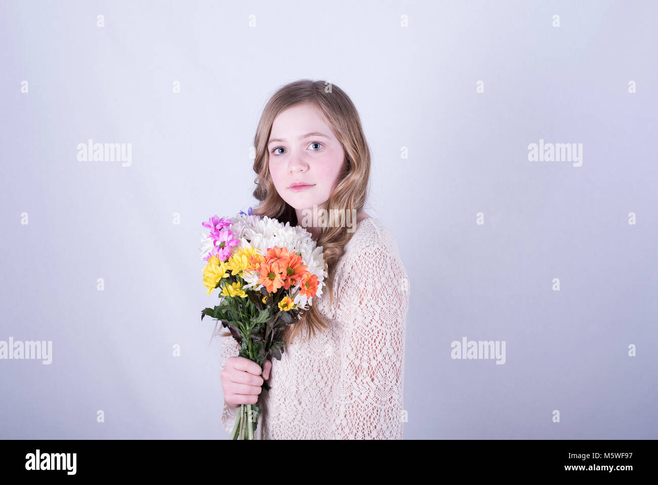 Jeune fille de 12 ans avec de longs cheveux blond sale incliné holding bouquet de marguerites colorées, les yeux grands ouverts, l'expression triste, fond blanc Banque D'Images