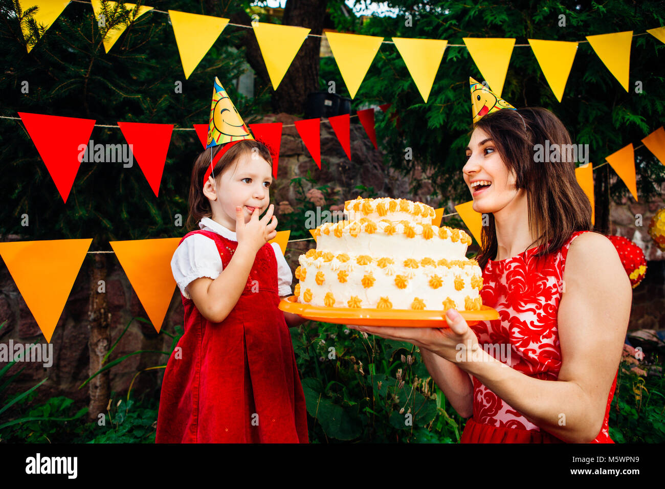 Fête pour les enfants et des gâteaux. Un petit enfant d'un funny girl tente de fabricants un grand gâteau de deux étages près de maman qui tient dans ses mains. Contre l'arrière Banque D'Images