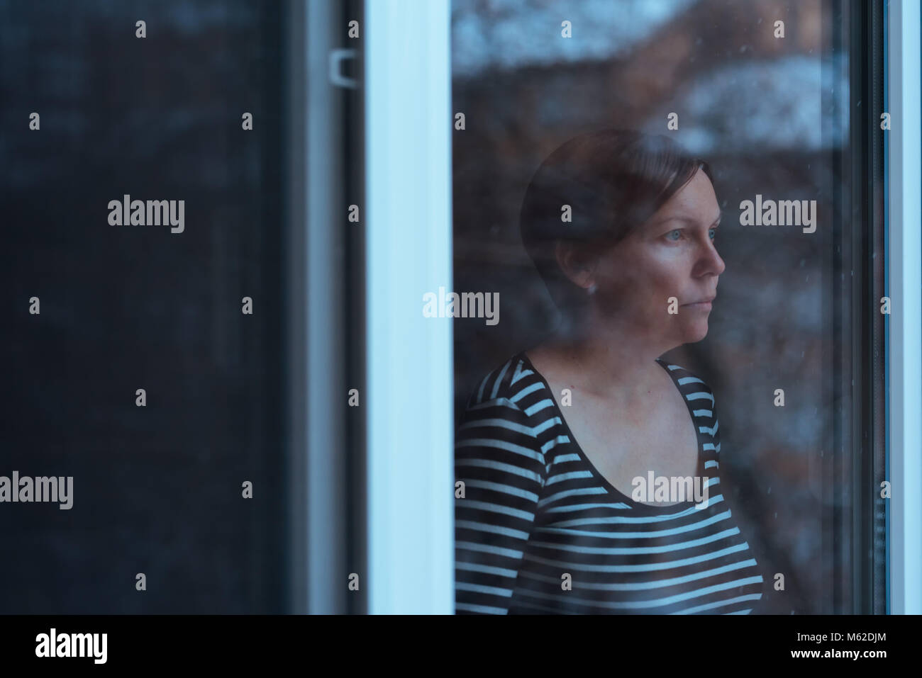 Femme debout appuyée par la fenêtre et à l'extérieur de la neige qui tombe en hiver froid après-midi, sarcelle et droit aux tons orange Banque D'Images