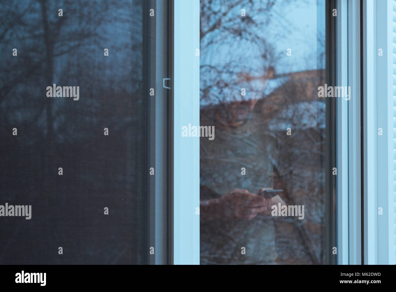 Seul homme triste à l'aide de téléphone mobile pour envoyer des sms par la fenêtre en hiver après midi alors qu'il neige à l'extérieur, Teal et tons orange avec image Banque D'Images
