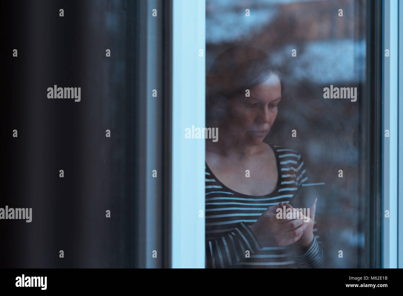 Seul homme triste à l'aide de téléphone mobile pour envoyer des sms par la fenêtre en hiver après midi alors qu'il neige à l'extérieur, Teal et tons orange avec image Banque D'Images