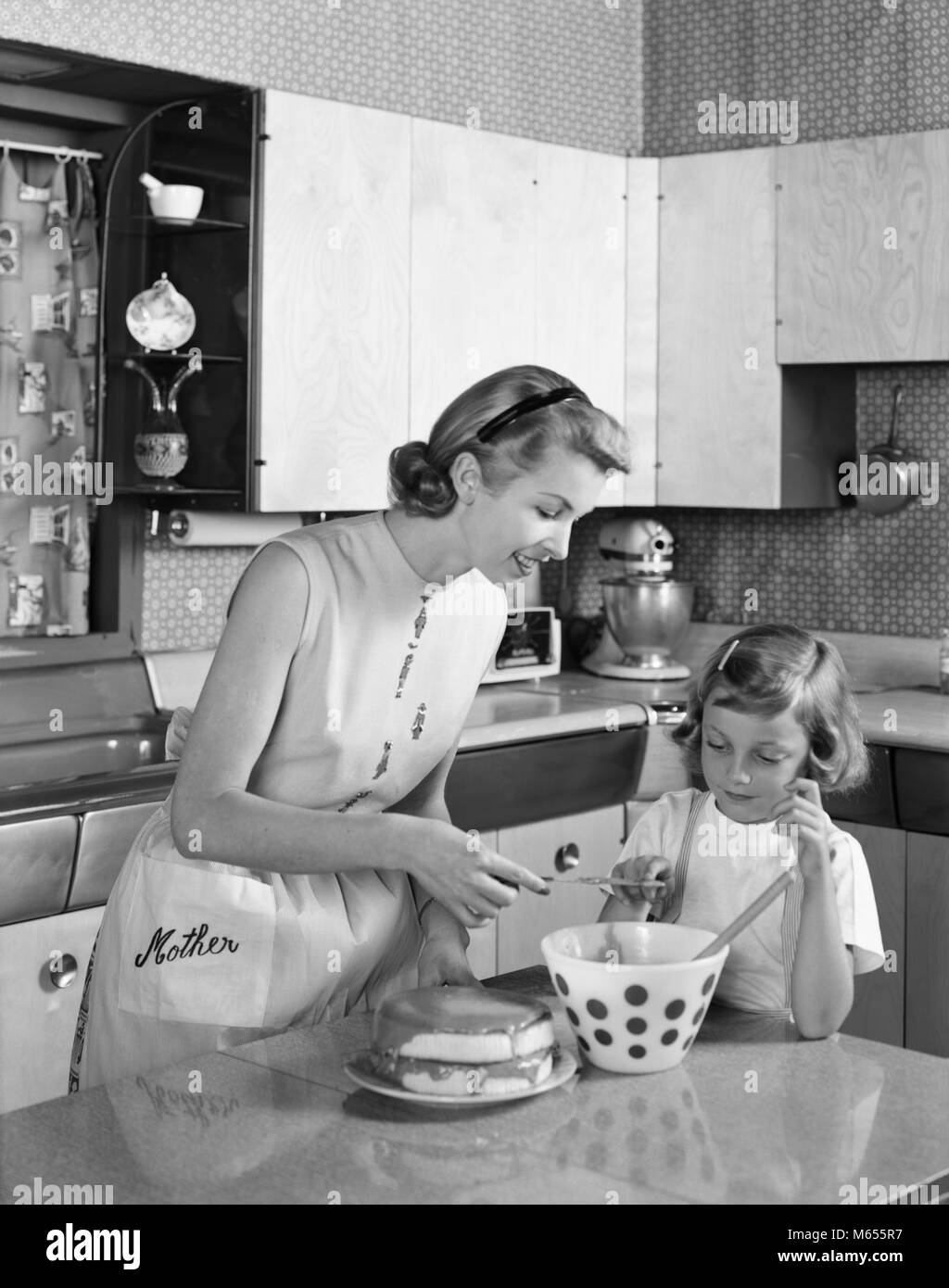 1950 Mère et fille EN GLAÇAGE GÂTEAU CUISINE DOUBLE COUCHE MOT MÈRE SUR TABLIER - asp x16750 CAM001 HARS, filles à l'intérieur des bols nostalgie de givrage AU FOYER ET LES MÈRES AU FOYER DE LA COUCHE DE GIVRE HOUSEWIVES MOT BOL JUVÉNILES B&W NOIR ET BLANC DE L'origine ethnique caucasienne Cuisine île old fashioned personnes montrant de l'enseignement Banque D'Images