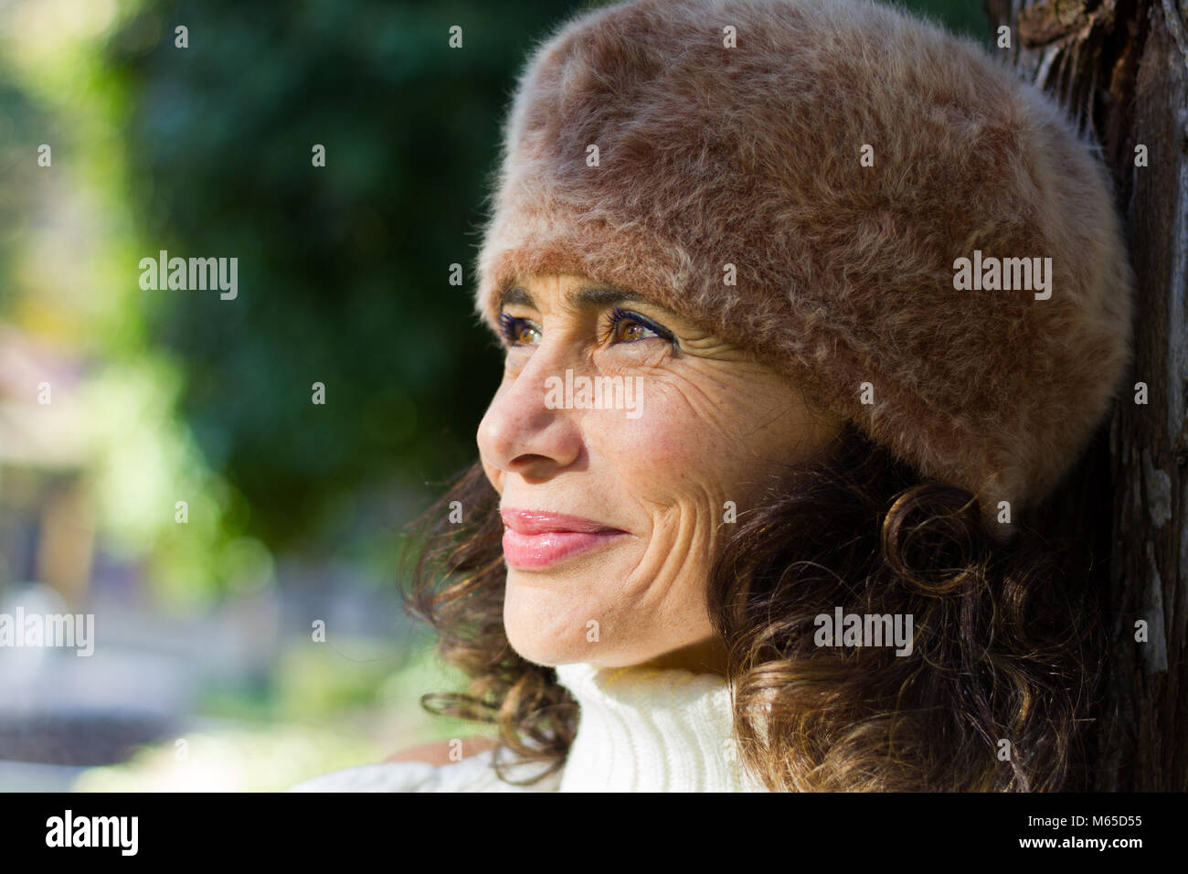 Bright close up portrait of young woman wearing éblouissante fourrure poilue beret hat and leaning on tree in park. Belle Dame d'âge moyen smiling portrait Banque D'Images