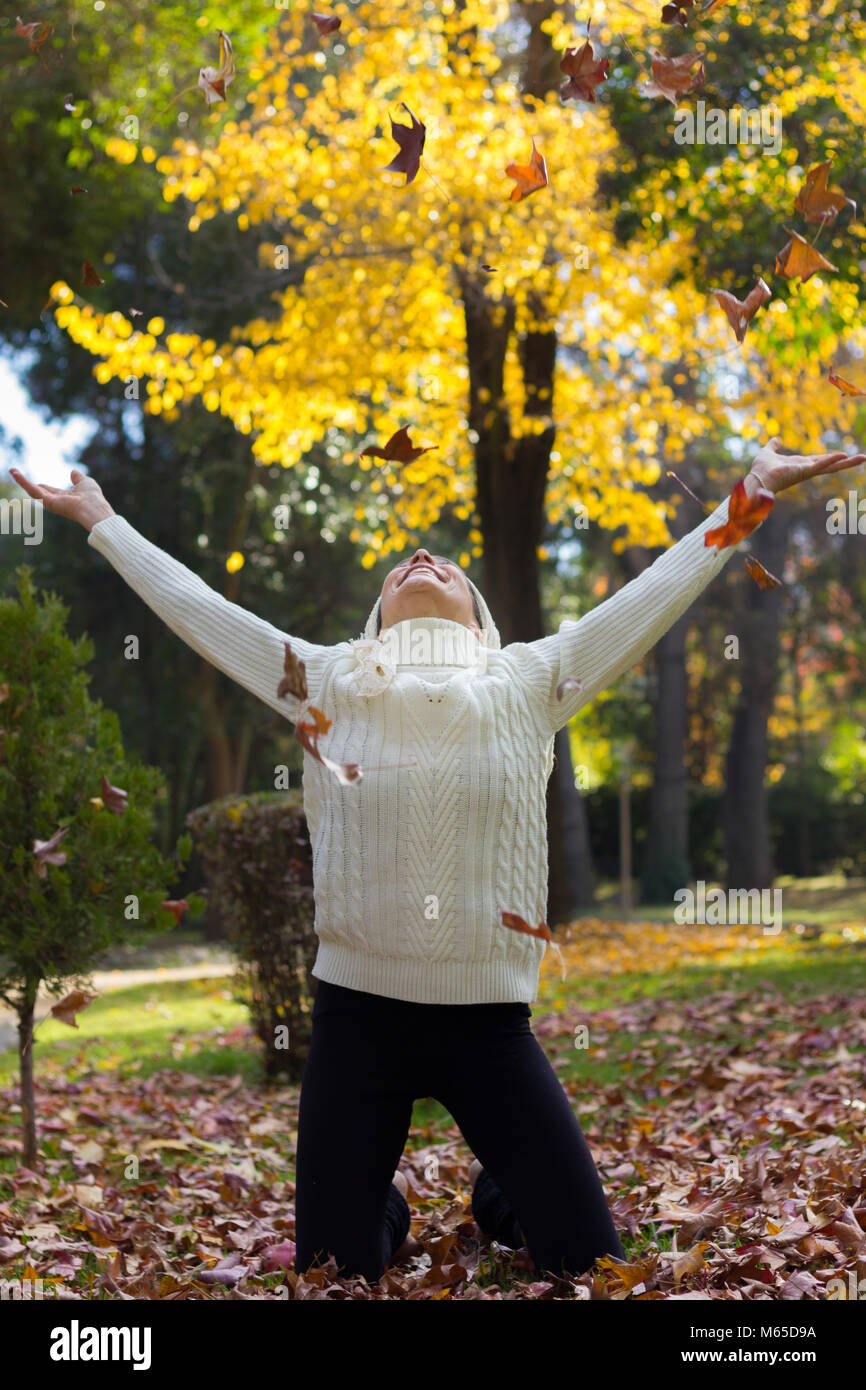 Femme euphorique se dresse sur ses genoux avec les bras ouverts à la recherche jusqu'alors que brown feuilles flottent dans l'air pendant l'automne dans le parc. Saison d'automne concept Banque D'Images