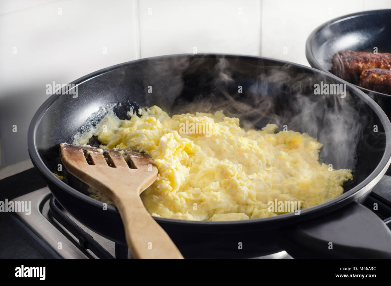 Des œufs brouillés et des saucisses végétariennes, la cuisson dans une poêle à frire ou noir sur le dessus de cuisinière avec spatule en bois et l'augmentation de la fumée. Mur carrelé blanc en background Banque D'Images